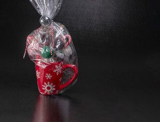 Wrapped red Christmas mug with snowflakes filled with cake pops, candy canes and marshmellows on a black background with text space.  Christmas gifts, baked gifts
