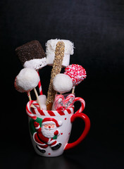 White Christmas mug with a Santa Claus picture filled with cake pops, marshmallows and candy canes on a black background with text space. Christmas baked gifts.