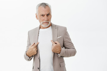 Portrait of senior man being accused in commiting crime standing insulted and intense pointing at himself with serious displeased and offended expression asking question at camera over gray wall