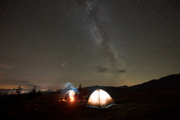 Night camping under beautiful starry sky. Group of four people, men and woman resting by burning campfire at tourist tents under dark sky with bright sparkling stars and Milky Way constellation.