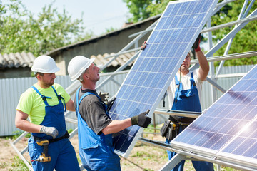 Team of three young technicians in protective helmets mounting heavy solar photo voltaic panel on...