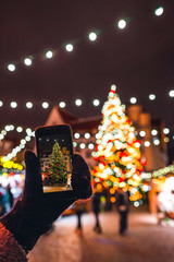 Hand holding phone with view on Christmas tree and Fair Market in Tallinn Old Town, traditional european Town Hall Square, loads of shining decoration, Estonia 