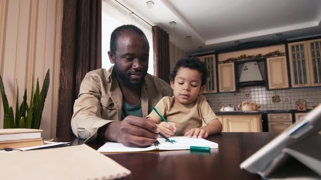 Panning shot of happy African American father sitting near his biracial child, imaging and drawing with felt pen in notebook at home