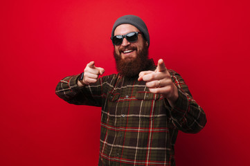 Photo of smiling  bearded man pointing and looking to camera over red background