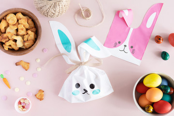 Top view and flat lay of Easter painted eggs, cookies, sprinkles and candies arranged on pink. Woman hands fill a bag or basket with sweet gifts