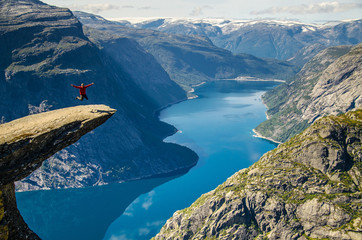 A man in a red jacket jumping on the Trolltunga rock with a blue lake 700 meters lower and interesting sky with clouds