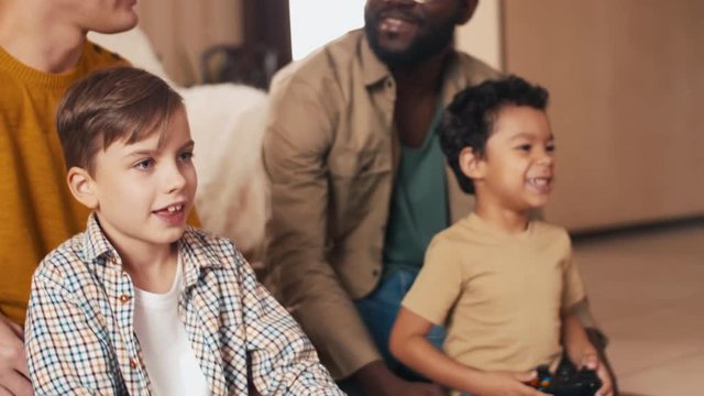 Two multi-ethnic friends discussing plans for weekend while their sons sitting near and using joysticks to play video games
