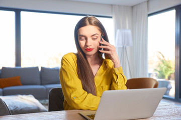 Young woman talking with somebody on her mobile phone and using laptop at home