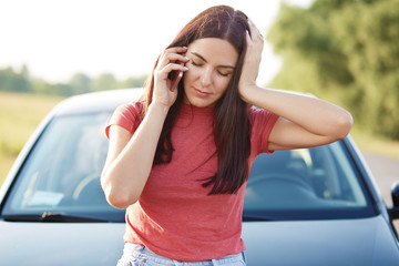 Tired European woman with long hair has telephone conversation via mobile phone, feels tired after repairing car, asks for help, dressed in casual t shirt. People, conversation, transport concept
