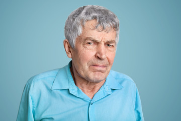 Close up shot of elderly man with wrinkles on face, pressed lips, looks scrupulously and with anger, discontent with something, dressed in blue shirt. Negative emotions and feelings concept.