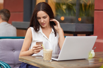 Horizontal shot of busy female freelancer holds mobile phone, text messages online, uses laptop computer, drinks milkshake, poses in outdoor restaurant, dressed in white clothes, sits in front of pc