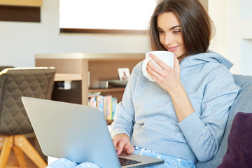 Young woman with her laptop sitting on sofa at home and drinking tea