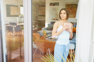 Beautiful young woman looking away while standing behind the window and drinking tea