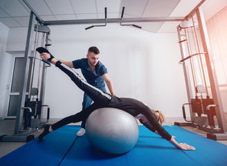 Concept of rehabilitation . Beautiful young woman doing exercises with ball under supervision of...