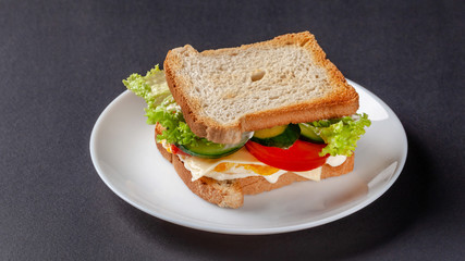 Homemade sandwich made of toast bread, fried egg, melted cheese, tomatoes, cucumber, lettuce, burgers and mayonnaise sauce. Sandwich on a white plate, on a black background.