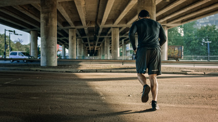 Backshot of an Athletic Young Man in Sports Outfit Jogging in the Street. He is Running in an Urban Environment Under a bridge with Cars in the Background.