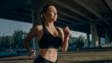 Fototapeta na wymiar Close Up Shot of a Beautiful Busty Fitness Girl in Black Athletic Top Jogging in a Sunny Street. She is Running in an Urban Environment Under a Bridge with Cars in the Background.