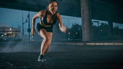 Fototapeta na wymiar Beautiful Strong Fitness Girl in Black Athletic Top and Shorts Starts Sprinting. She is Running in an Urban Environment Under a Bridge with Cars in the Background.