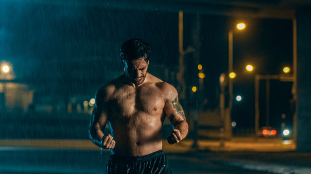 Shirtless Athletic Muscular Young Man is Celebrating His Sport Accomplishments on a Rainy Night and Showing His Muscles. He is in an Urban Environment Under a bridge with Cars in the Background.