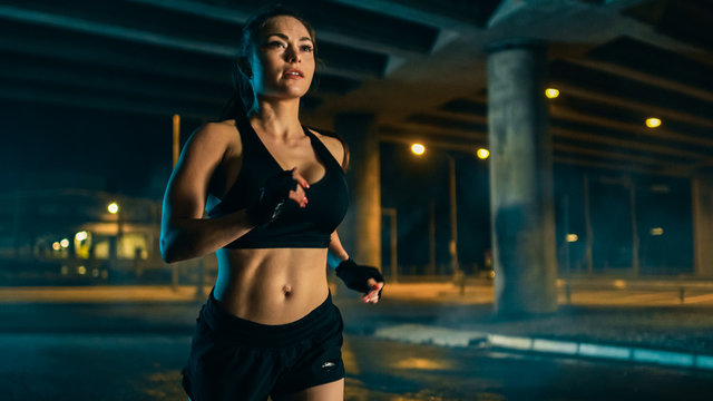 Beautiful Busty Fitness Girl in Black Athletic Top and Shorts is Jogging on the Street. She is Doing a Workout in a Night Urban Environment Under a Bridge.