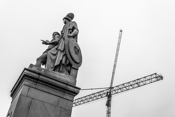 Zwei Statuen auf einem Sockel vor zwei Baukränen - monochrom