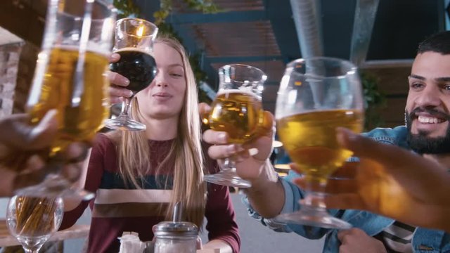 Track in shot of young Caucasian woman hanging out with friends in pub, proposing a toast and clinking glass of beer