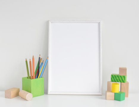 Blank frame in kids room, white frame mock up for prints, artwork, photos or painting, colorful pencils and building blocks.