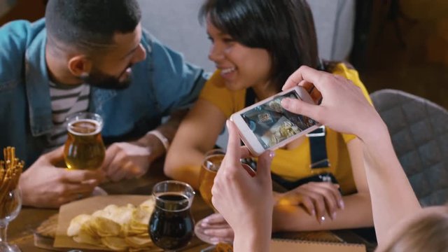 Tilt up shot of Caucasian woman snapping a picture of table settings while her friends talking and laughing at the table with beer and snacks inside the bar