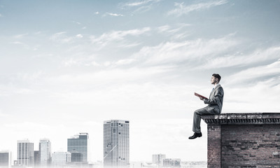 Man on roof edge reading book and cityscape at background