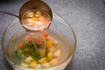 soup in a glass bowl is poured by a scoop. broccoli, carrots, potatoes, chicken