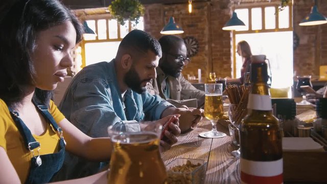 Close up of young people sitting at bar counter with glasses of beer and checking social networks with their telephones