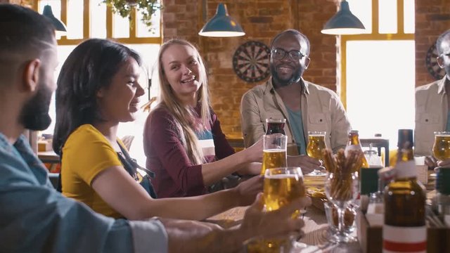 Multiracial friends congratulating Latin American woman with her birthday, clinking glasses of beer and drinking it inside the pub