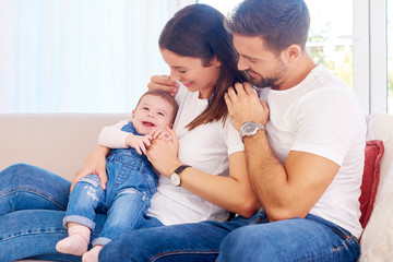 Cheerful young family with their cute baby girl sitting on sofa