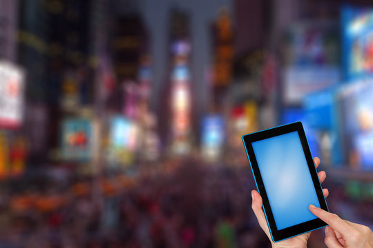 Female finger touching tablet blue blank touchscreen ready for your text. Intentionally blurred image of a night Times Square (NYC) is in the background. All potential trademarks are removed.
