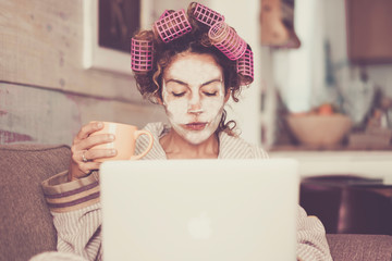Rare and funny portrait of beautiful woman with facial mask and curlers hair taking a coffee at home while use internet with modern laptop computer - home scene for modern lifestyle people 