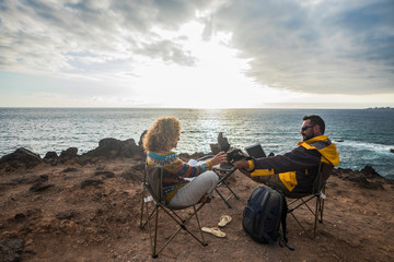 Couple of alternative tourists traveler sit down with the ocean in front and enjoy the rest and the landscape working at laptop connected computer or reading a paper book - sunset and nature 
