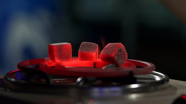 Kindling of square coal for a hookah on a special furnace with a hot spiral. Close-up of kindling coals for hookah in hookah bar. Man holds red-hot coal with tongs.