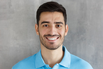 Close up portrait of young smiling handsome man in blue polo shirt, standing against gray textured wall