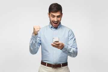Portrait of excited young business man looking at smart phone isolated on gray background. Winner...