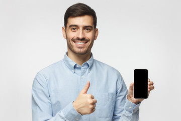 Happy young man holding blank smartphone, smiling at camera, showing thubms up gesture, isolated on gray background