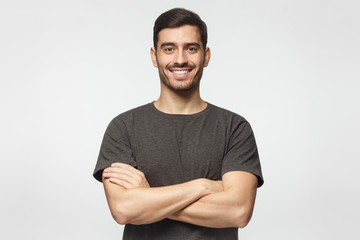 Smiling handsome young man in grey t-shirt standing with crossed arms, isolated on gray background