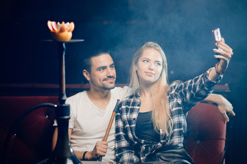 Beautiful couple in nightclub smoking hookah, girl take selfie on smartphone