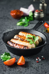 Grilled sausages in cast iron skillet with herbs, sauce and spices on dark background. Selective focus, space for text.