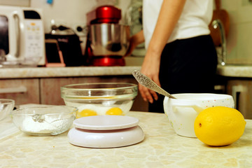 A woman is cooking in her kitchen, about to bake a cake. In the foreground - small kitchen scales, lemon, sugar bowl, a glass bowl with egg yolks