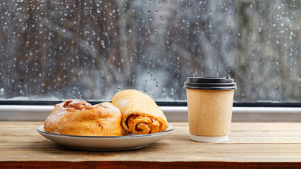 Fresh pastry and paper cup of coffee on vintage wooden windowsill or table against window with raindrops on blurred background. Shallow focus.