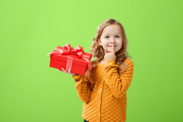 Portrait of a little girl in a warm sweater on a ufo green background. The child looks dreamily up and holds a red box with a gift. The concept of celebration, giving and receiving a gift