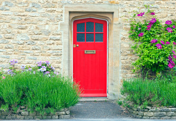 Fototapeta na wymiar Bright red wooden doors in an old traditional English stone house, surrounded by climbing pink clematis on the golden wall, lavender in the front garden .