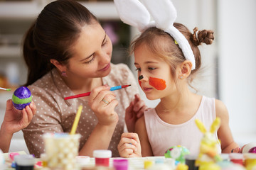 Mother draws on daughter's face while dyeing the eggs for the Easter table in the cozy light kitchen.