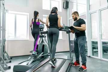 Tuinposter Young smiling fitness women with personal trainer an adult athletic man on treadmill in the gym. Sport, teamwork, training, healthy lifestyle concept © Valerii Honcharuk
