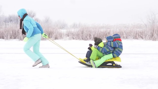 Mom in blue ski suit and three happy children in warm winter clothes ride on yellow sled on frozen snow-covered river.  Concept of friendly sports family. Winter holidays in village. Christmas eve